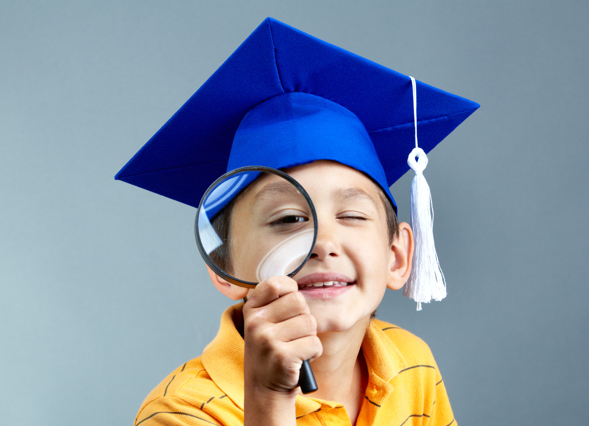 Portrait of curious boy in graduation hat looking through magnifying glass